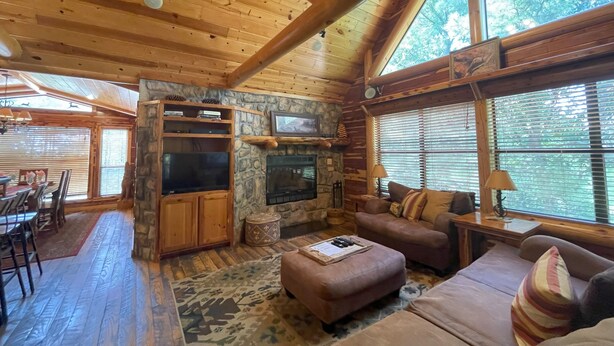 L’intérieur chaleureux d’un chalet avec une cheminée en pierre, des poutres en bois au plafond, un coin repas et de grandes fenêtres offrant une vue sur l’extérieur.