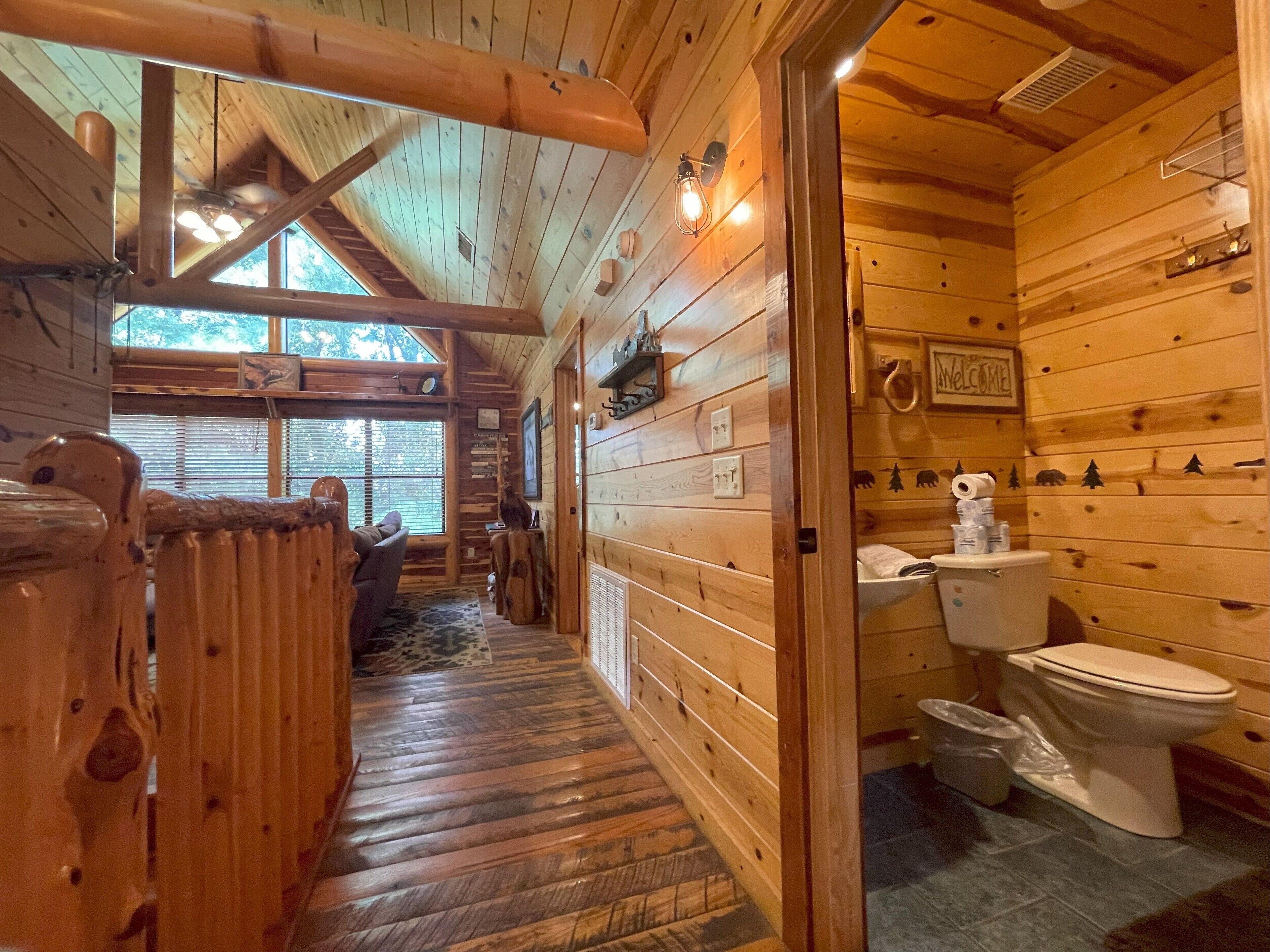 A wooden cabin interior with a wooden floor, a dining area with a table and chairs, a wooden ceiling with exposed beams, and a bathroom with a toilet and sink.