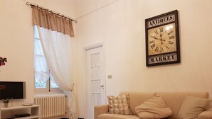 Ein Wohnzimmer mit beiger Couch, einem Fernseher auf einem Schrank, einer Heizung und einem Fenster mit durchsichtigen Vorhängen.