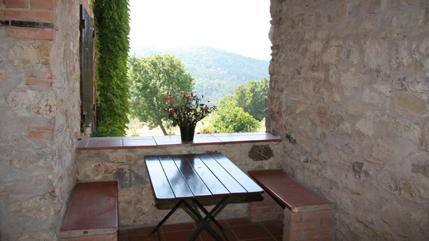 A rustic stone wall with a wooden table and bench, a potted plant with red flowers, and a view of green hills. - Tuscan holiday home near the sea between Siena, Florence and Pisa (Montieri)
