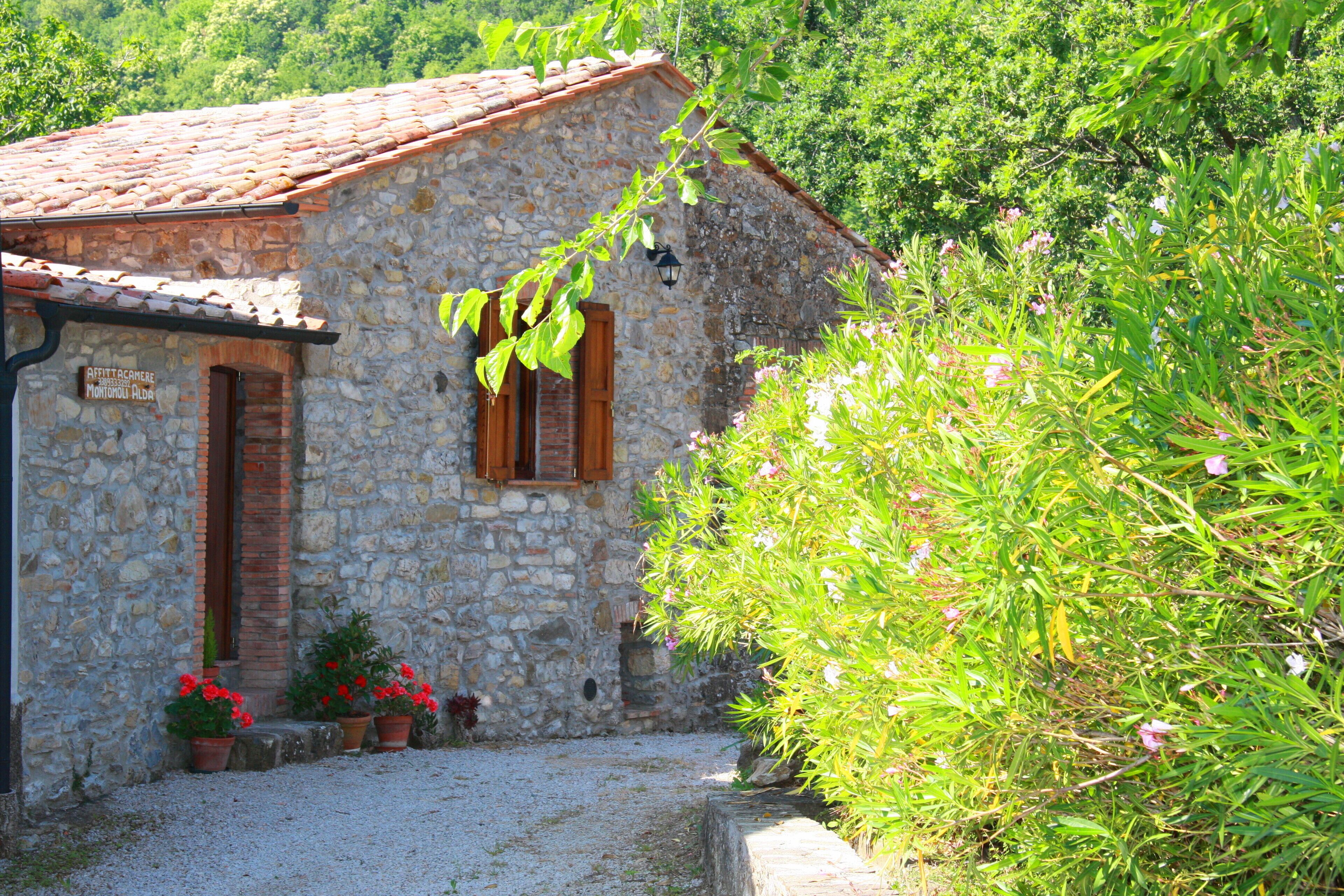 Il s’agit d’un bâtiment en pierre, doté d’un toit en tuiles, d’une porte en bois et d’une fenêtre avec des volets. Devant le bâtiment, on peut voir des plantes en pot ornées de fleurs rouges.