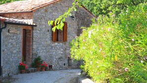A stone building with a tiled roof, a wooden door, and a window with shutters. There are potted plants with red flowers in front of the building. - Tuscan holiday home near the sea between Siena, Florence and Pisa (Montieri)