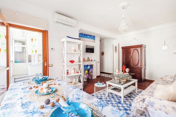 Una habitación bien iluminada con una mesa de comedor lista para una comida, un estante blanco con adornos, un mueble de madera y un televisor en la pared.