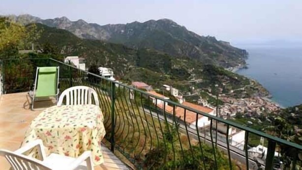 A balcony with a table and chairs, overlooking a coastal landscape with mountains and a view of the sea. - RAVELLO "Nonna Anna" AMALFI COAST (RAVELLO)
