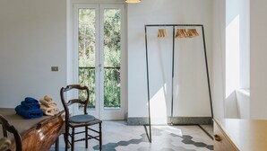 Een kamer met een houten stoel, een tafel met handdoeken erover gevouwen, een zwarte vloerlamp, en uitzicht door een glazen deur op groen.