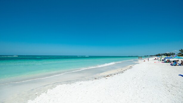 A beach with clear turquoise water, white sand, and a few people and umbrellas on the shore.