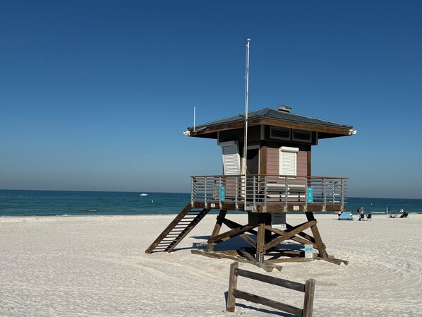 Torre de salvavidas con estructura de madera, barandillas metálicas y asta de bandera en una playa de arena bajo un cielo azul despejado.