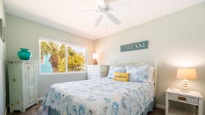 A bedroom with a bed, a nightstand, a dresser, and a window with a view of palm trees.