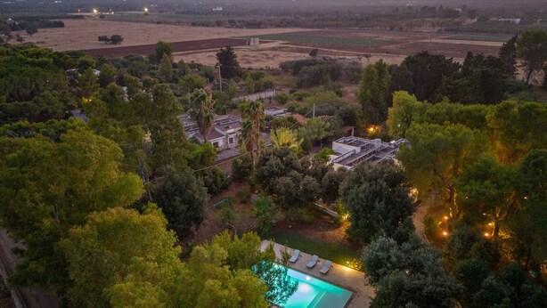 Vista aérea de um bairro residencial com piscina cercada por árvores, uma casa com telhado de cerâmica e campos abertos ao fundo.