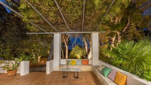 Um pátio externo com mesa e assentos, cercado por plantas e árvores.
