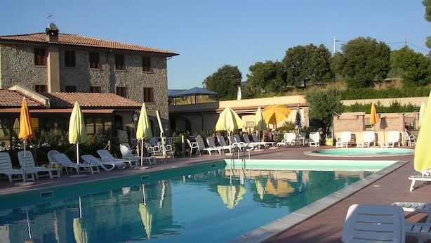Zona de piscina con sillones de descanso y sombrillas, un edificio de piedra al fondo y cielo despejado.