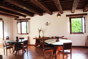 Zona pranzo con tavolo rotondo e sedie, travi in legno al soffitto e finestre che si affacciano sul verde.