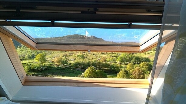 A skylight with a view of a green landscape and a distant wind turbine. - The Pyrenees footbridge (Unac)