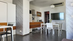 Une cuisine moderne avec des meubles blancs, un coin repas avec une table et des chaises blanches, et un salon avec un canapé et un téléviseur fixé au mur.