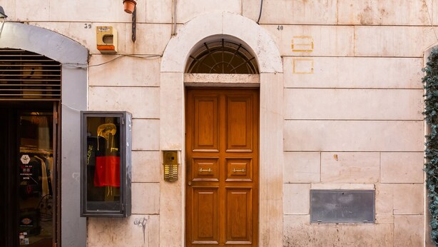 Une façade d’immeuble avec une porte double en bois, une entrée de magasin avec une vitrine, et une petite plaque fixée au mur.
