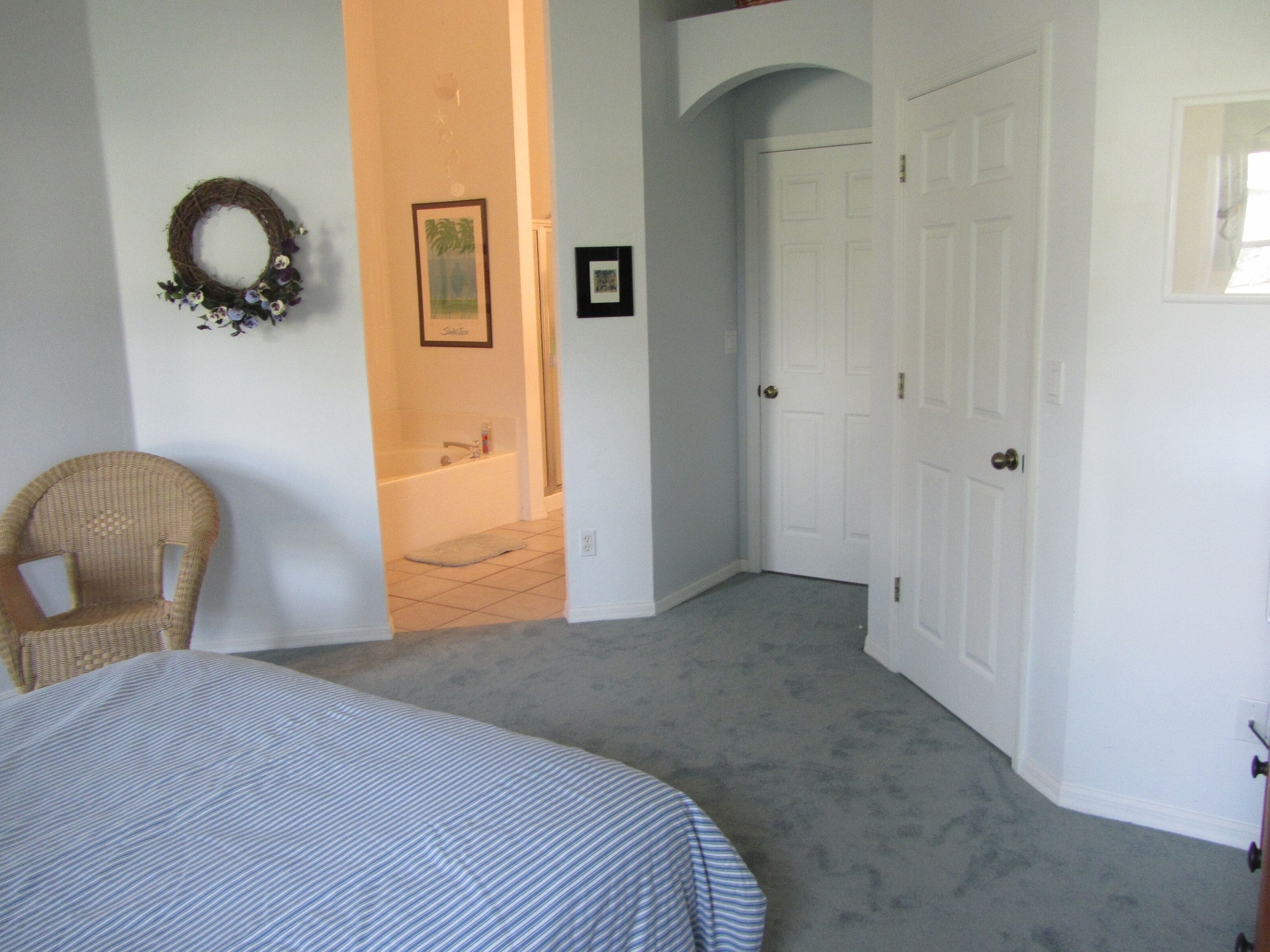 A bedroom with a bed, a wicker chair, a bathroom with a bathtub, and a door leading to another room.