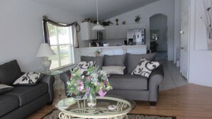A living room with a grey sofa, a glass coffee table with a floral centerpiece, and a kitchen in the background.