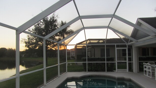A covered outdoor swimming pool with a glass enclosure, surrounded by a lawn and a body of water.