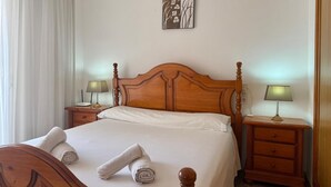 Un letto rifatto con lenzuola bianche e due asciugamani arrotolati, comodini in legno con lampade, un quadro appeso al muro e una finestra con tende bianche.