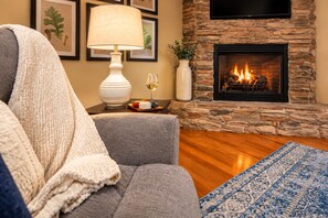 A cozy living room with a stone fireplace, a comfortable gray sofa, a white lamp, and a blue patterned rug. - Private Hot Tub, Quiet Setting, Spotlessly Clean Cottage (Asheville)