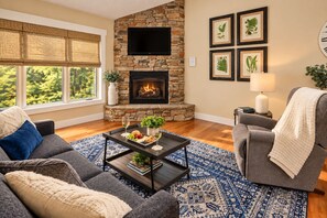 A living room with a stone fireplace, a flat-screen TV, and framed botanical prints on the wall.