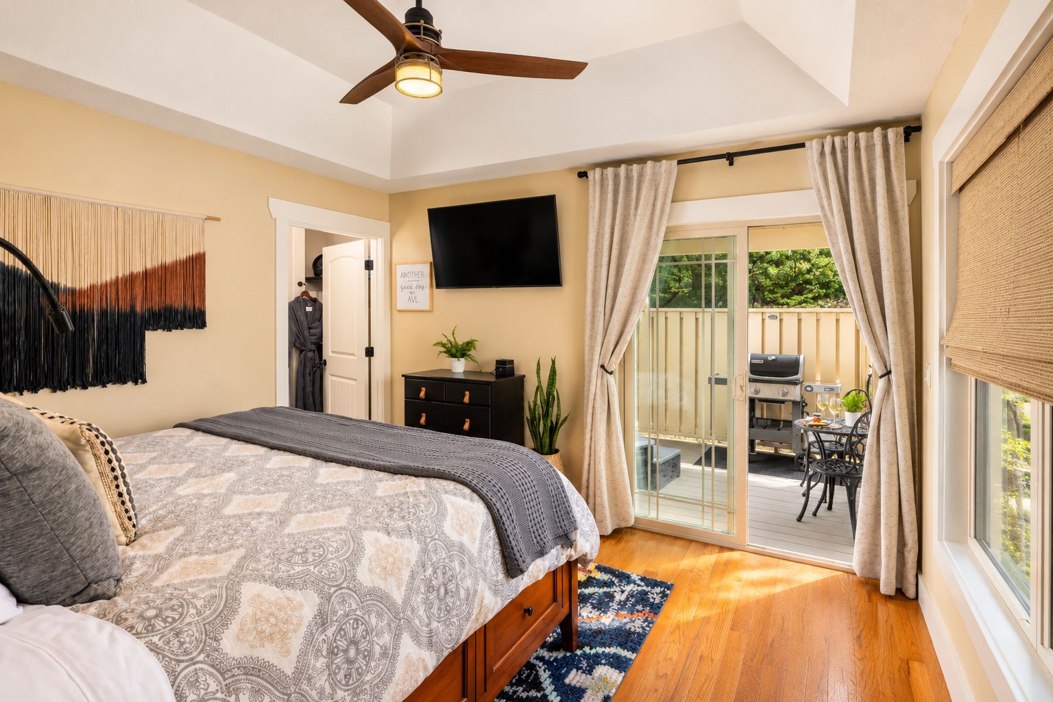 Ein Schlafzimmer mit Bett, Deckenventilator, Fernseher und einer Schiebetür zu einer Terrasse im Freien mit Grill und Sitzgelegenheit.