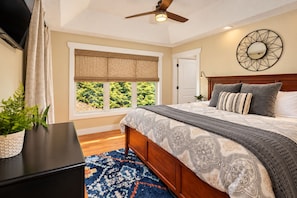 Quarto com cama, ventilador de teto, janela vista para vegetação e espelho fixado na parede.