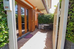 Een overdekt terras buiten met een tafel en stoelen, een houten deur en een kleine tuin.