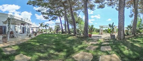 Un jardín bien cuidado con senderos de piedra, árboles grandes y un edificio blanco con mobiliario exterior.