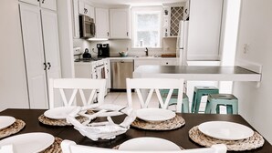 Une cuisine moderne avec des meubles blancs, un îlot central et un coin repas agrémenté de chaises blanches et de sets de table en fibres tressées.