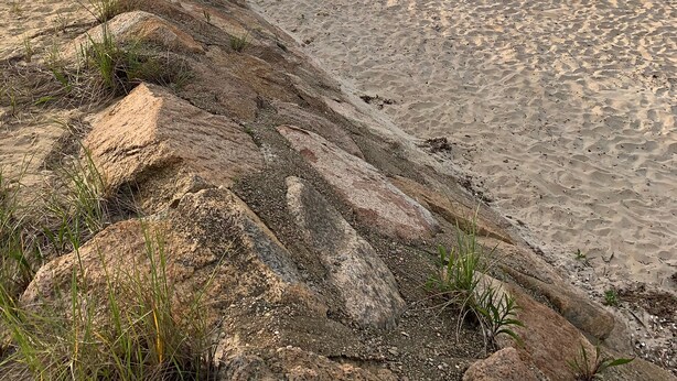 Une plage avec un escalier en bois descendant jusqu’au rivage, un promontoire rocheux et une mer calme.