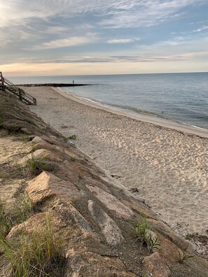 A beach with a wooden staircase leading down to the shore, a rocky outcrop with sparse vegetation, and a calm sea.