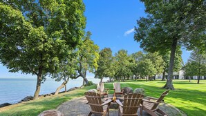 A circular outdoor seating area with Adirondack chairs and a fire pit, surrounded by a brick patio and large rocks, overlooking a body of water and a well-maintained lawn. - Izaty's Beach Villa- Completely remodeled and exceptionally clean (Onamia)