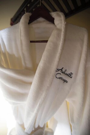A white robe hanging on a wooden hanger with the text "Ashville Cottages" embroidered on it.