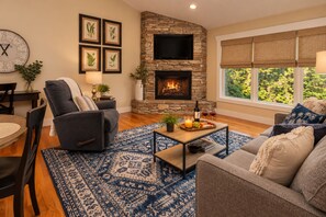 A living room with a stone fireplace, a clock, framed botanical prints, and a dining area with a table and chairs.