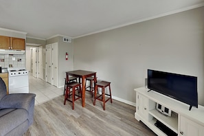 A compact living space with a kitchenette, a dining area with a table and chairs, a television on a stand, and a fire extinguisher mounted on the wall. - Beach Racquet A223 | Partial Beach Views + Pool (Tybee Island)