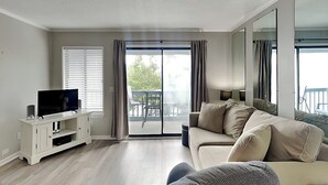 A living room with a flat-screen TV, a sofa, a coffee table, and a sliding glass door leading to an outdoor area with a table and chairs.