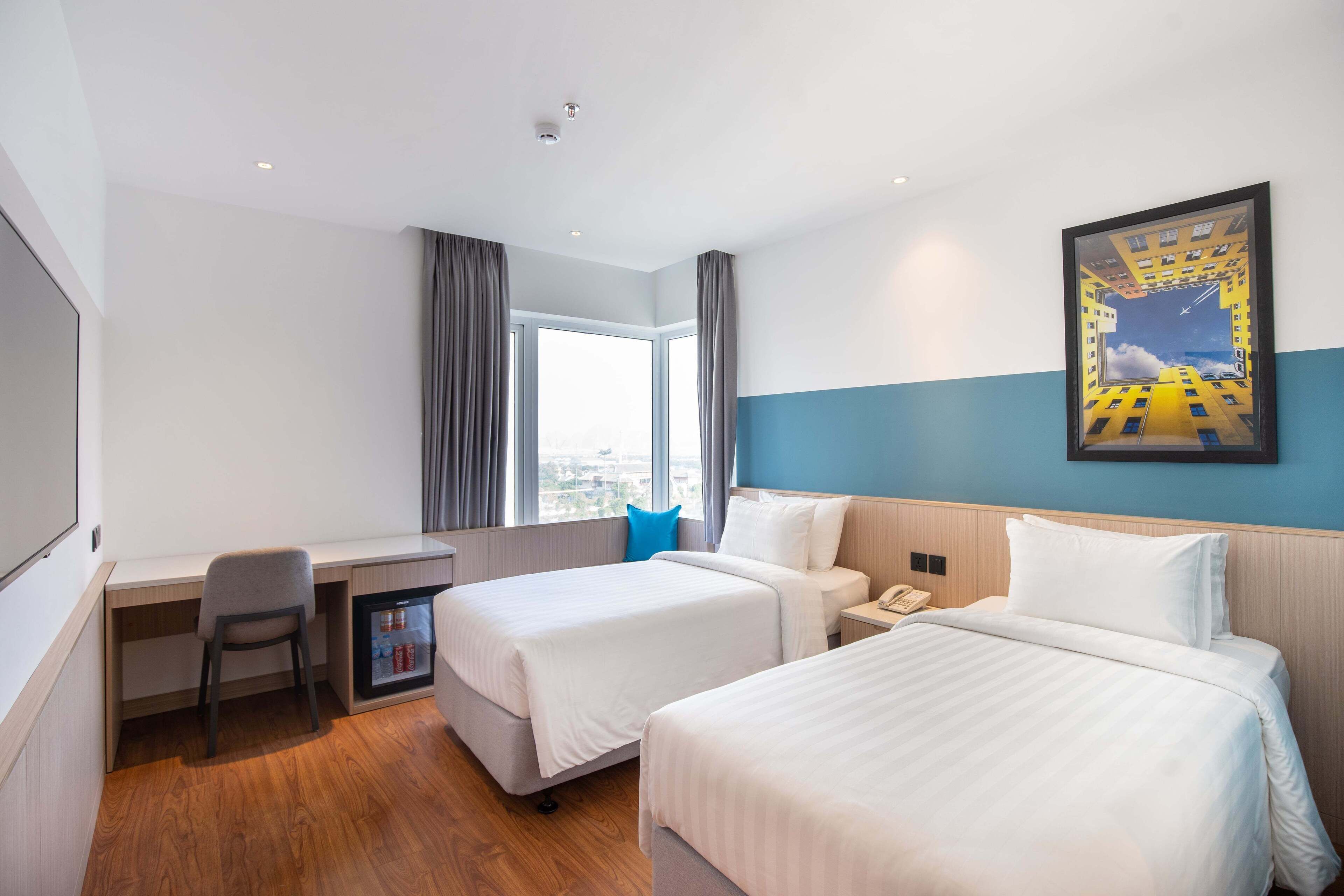 A hotel room with two beds, a desk, a chair, a small refrigerator, and a window with curtains.
