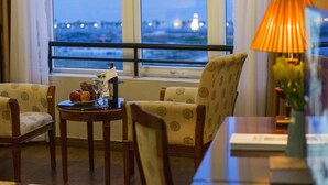 A room with two patterned armchairs, a small table with a bottle and fruit, and a view of a body of water through large windows. - Halong Pearl Hotel (Bai Chay)
