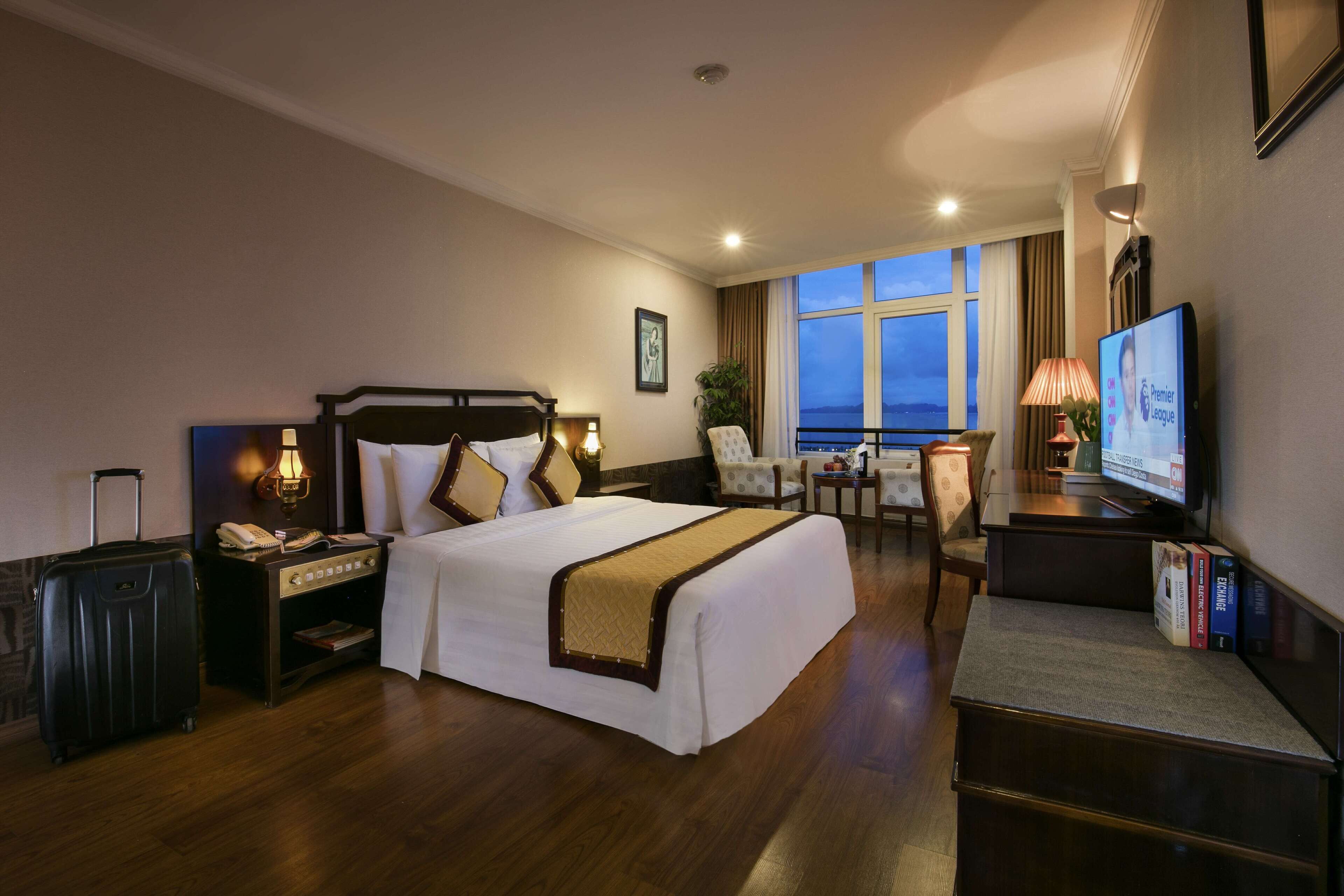 A hotel room with a large bed, bedside table with a lamp, a desk with a chair, a television, and a view of the outside.
