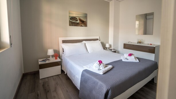 A hotel room with a large bed, two bedside tables, a nightstand, a wall-mounted mirror, and a painting of a boat on the wall. - Anchor Apartment (Marina di Ragusa)
