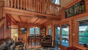 A wooden cabin interior with a 'WELCOME' sign, a dining area, a leather sofa, and a large window overlooking a deck. - Nice Views, Lake Access, 2 Kayaks, Log Home, Paved Roads - Honey Bee Hill (Lake Lure)