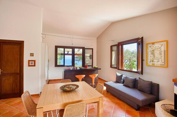 Un comedor con una mesa y sillas de madera, un sofá gris y una encimera de cocina con vistas al exterior.
