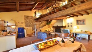 A spacious living area with a stone fireplace, a dining table set with fruits, and a kitchen in the background.