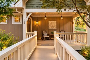 A wooden deck with a white railing, a seating area with chairs and a table, and a wall-mounted light fixture. - Super Clean Cottage with Private Hot Tub and Fireplace (Asheville)