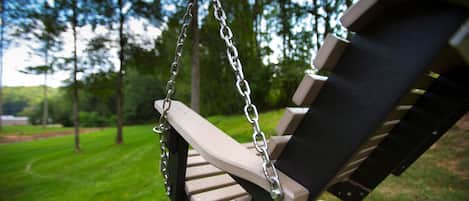A wooden swing set with metal chains, set against a backdrop of a grassy area with trees.