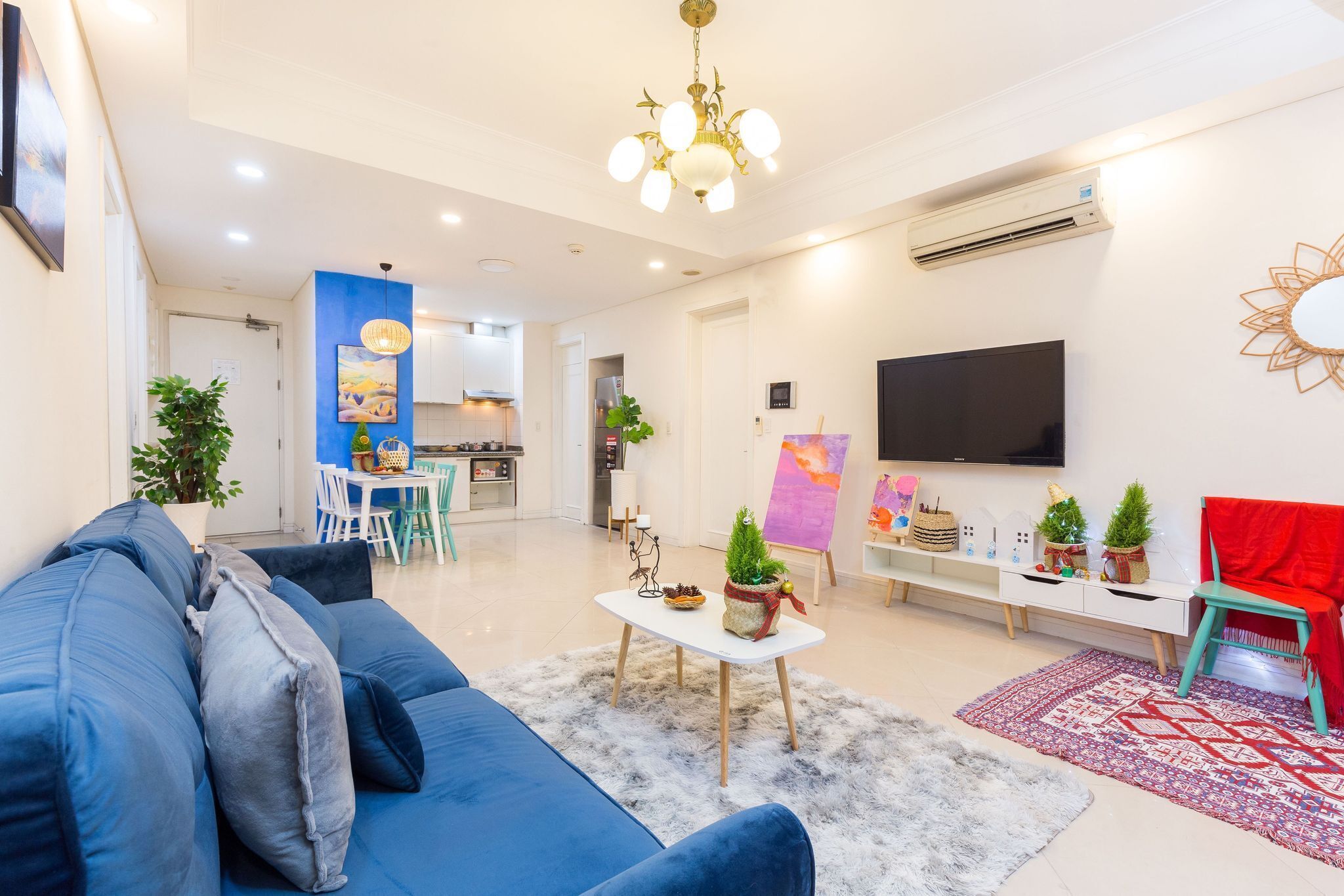 Uma sala de estar moderna com um sofá azul, uma mesa de centro branca e uma TV na parede. Cozinha visível ao fundo, com uma área de jantar.