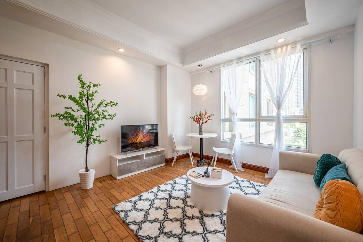 A modern living room with a flat-screen TV, a potted plant, a round coffee table, and a sofa with colorful cushions.