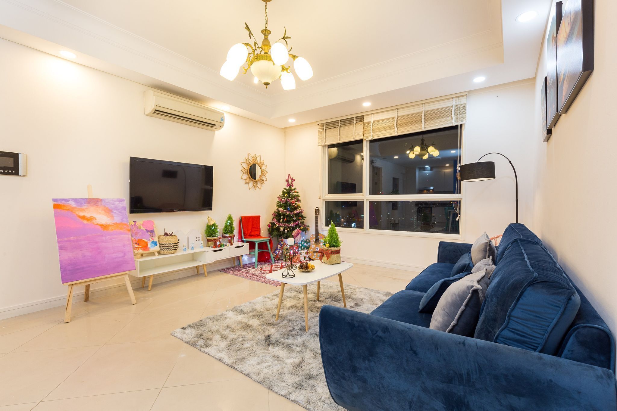 Ein Wohnzimmer mit einem blauen Sofa, einem Gemälde auf Staffeleien, einem Weihnachtsbaum und einem Fernseher an der Wand.