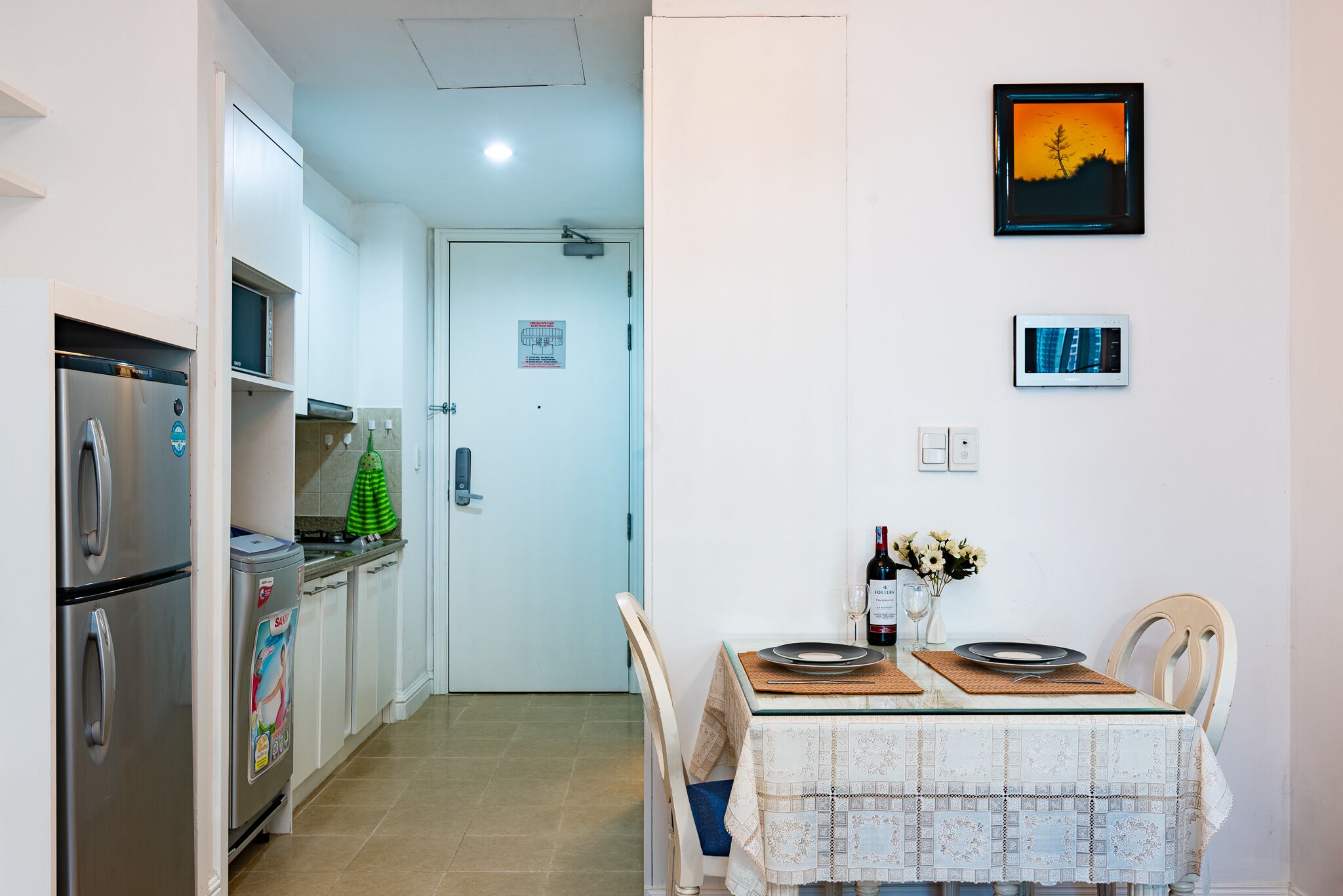 A compact kitchen with a refrigerator, microwave, and sink. A dining table with chairs is set against a wall with a framed picture.